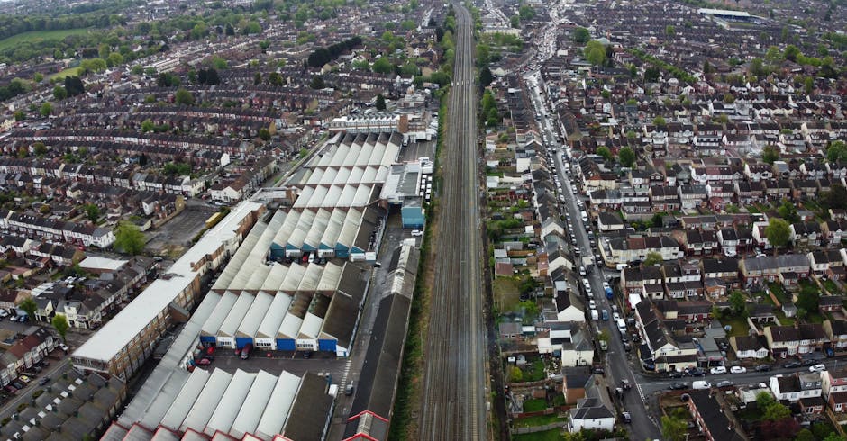 An aerial view showing a section of railway tracks running horizontally across the image, with a row of parked cars and vans positioned on a gravel and paved lot below. Several white and grey moving c