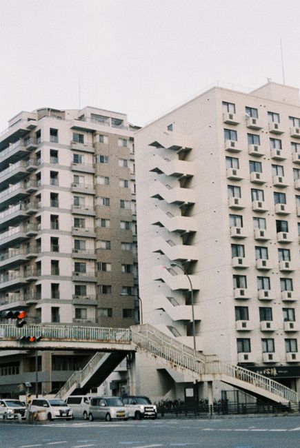 A street scene featuring two white multi-storey residential buildings with numerous small windows; the building on the right has external fire escape stairs that zigzag down its facade. In the foregro