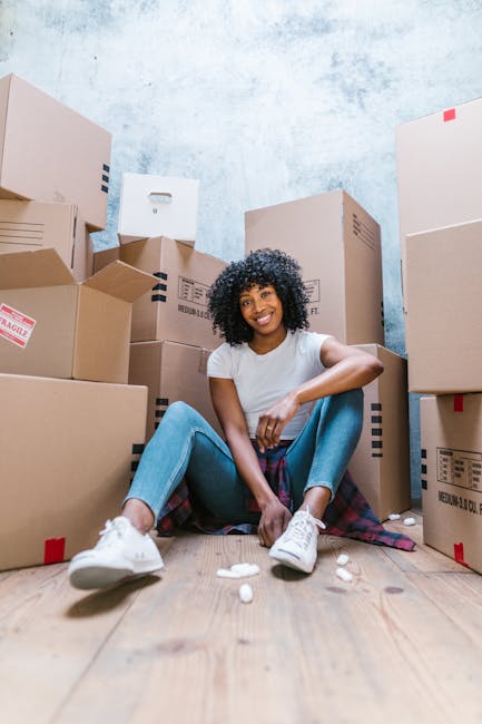 Two young women engaged in home relocation activities, standing among stacked cardboard moving boxes inside a room with a textured grey wall. The woman on the left holds a medium-sized box with a red 